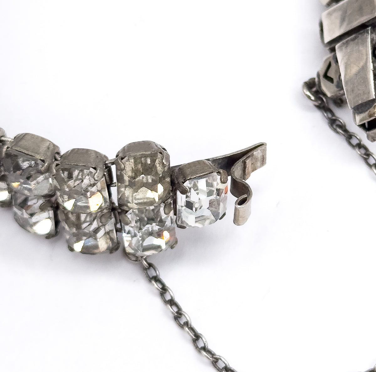 Macro of the open clasp end, its curled metal hook and tongue beside the last emerald cut stones, with the fine safety chain trailing away across the white surface.