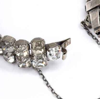 Macro of the open clasp end, its curled metal hook and tongue beside the last emerald cut stones, with the fine safety chain trailing away across the white surface.