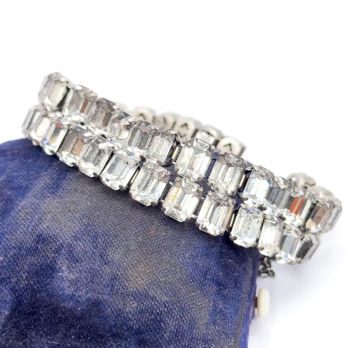 Art Deco bracelet of two rows of emerald cut colourless glass stones in prong-set white base metal, shown draped over a deep blue velvet cushion.