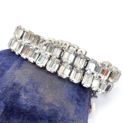 Art Deco bracelet of two rows of emerald cut colourless glass stones in prong-set white base metal, shown draped over a deep blue velvet cushion.