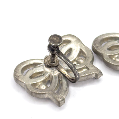 A closer look at the back of one earring with the second just entering the frame, the flat screw knob turned upward showing tiny stamped lettering above the curved wire arm.