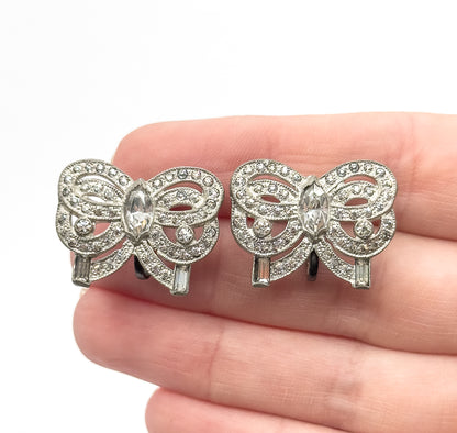A pair of rhinestone bow earrings in base metal, held on fingertips so their size reads small, each bow set with a marquise stone at the centre, round stones on the loops and baguettes at the ends.