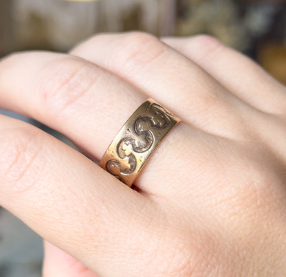 Victorian trefoil cigar band ring in rose gold, shown on a finger, the wide band's raised trefoil pattern curving across the face towards a small cartouche.
