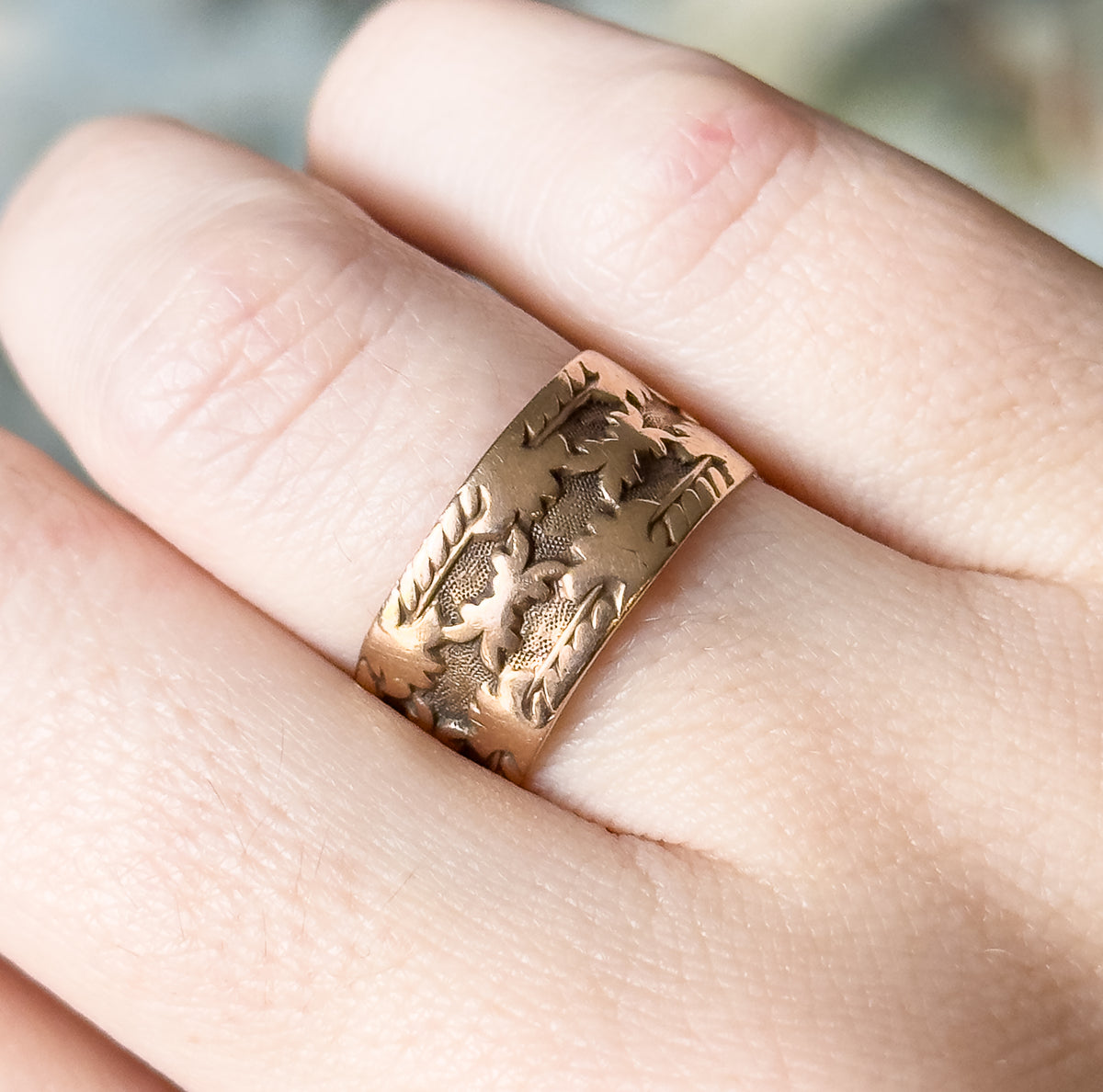 Overhead view of the band on the finger between the knuckles, its carved leaf surface catching the light and its width spanning much of the finger.