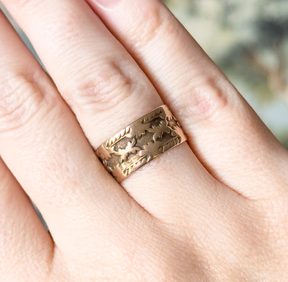 A Victorian wide rose gold band carved with leaf details around the shank, shown on a finger with the pattern facing the camera, giving a sense of its width.