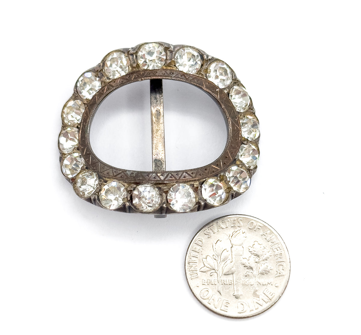 The buckle lies face up beside a dime, the coin reaching roughly half the height of the oval frame and its ring of colourless paste stones around the etched sterling silver.