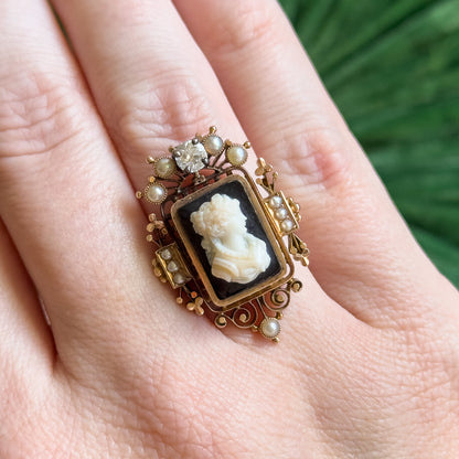 Shown on a finger against green foliage, the black and white cameo spans the full width between knuckles, seed pearls scattered around it and the prong-set diamond sitting at the top.