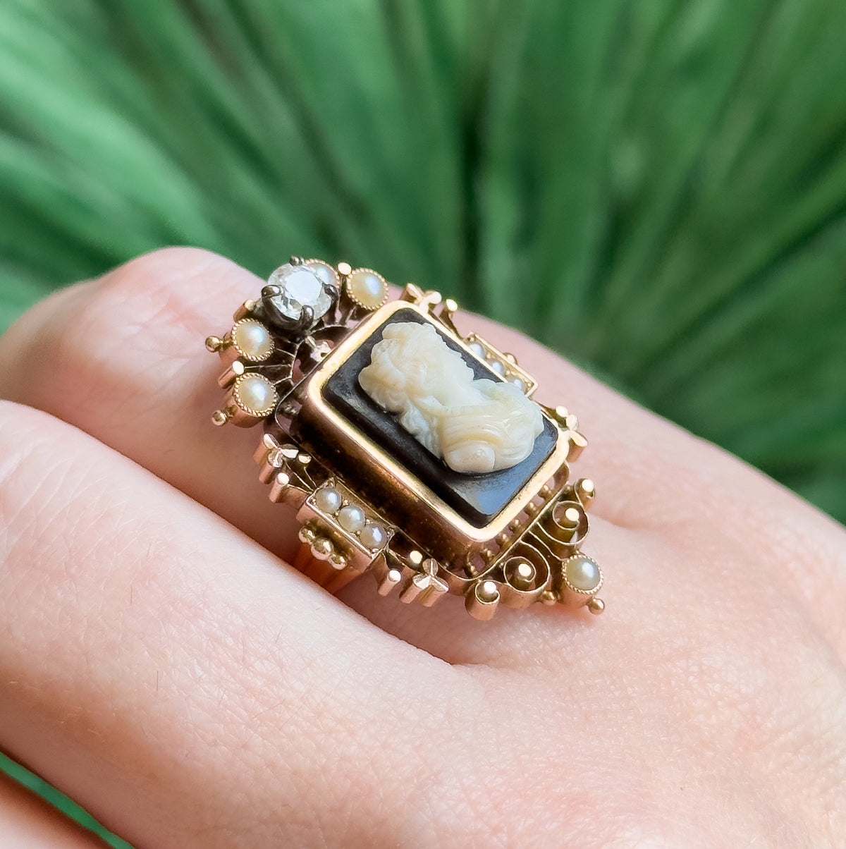 Close three-quarter view on the finger, the carved white figure standing proud of the black ground inside its rose gold bezel, with beaded scrollwork and seed pearls framing each side.
