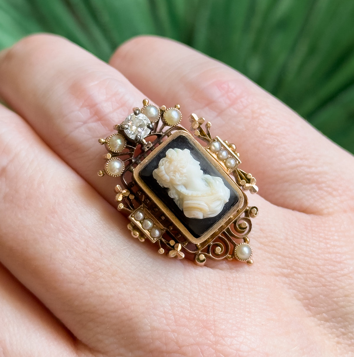 Shown on a finger with the setting tilted diagonally, the lozenge-shaped frame reaches from knuckle to knuckle, its scrolled lower point and pearl-set corners catching the light.