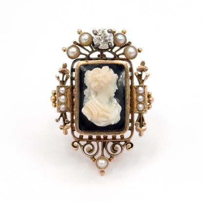 Straight-on frontal view against white, the whole face symmetrical: the prong-set diamond and fanned pearls above, the cameo centred, and a single pearl below the scrolled openwork base.