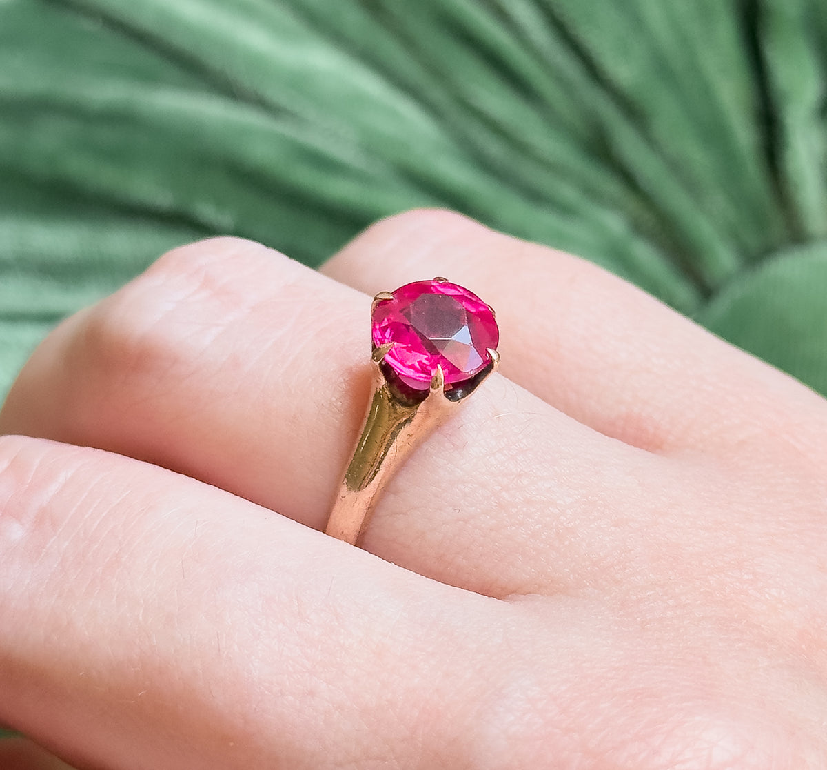 Shown on a finger against a soft green backdrop, the bright pink round stone sitting high in its prongs and reading large across the finger.