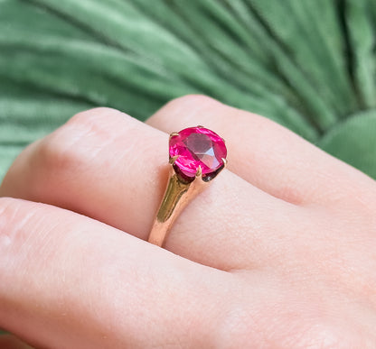 Shown on a finger against a soft green backdrop, the bright pink round stone sitting high in its prongs and reading large across the finger.