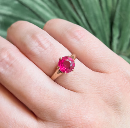 Closer view looking down onto the hand, the four prongs gripping the bright pink round stone clearly visible and the slim rose gold shank curving away across the finger.