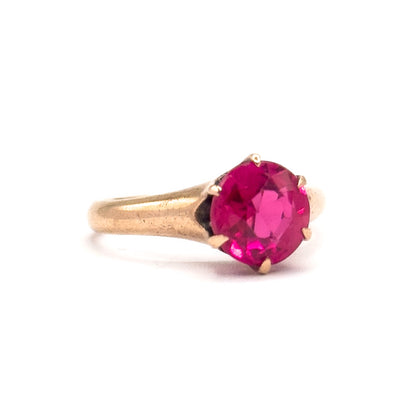 Edwardian synthetic ruby solitaire ring in 10K rose gold, shown in profile against white, the bright pink round stone held high in prongs above a tapering shank.