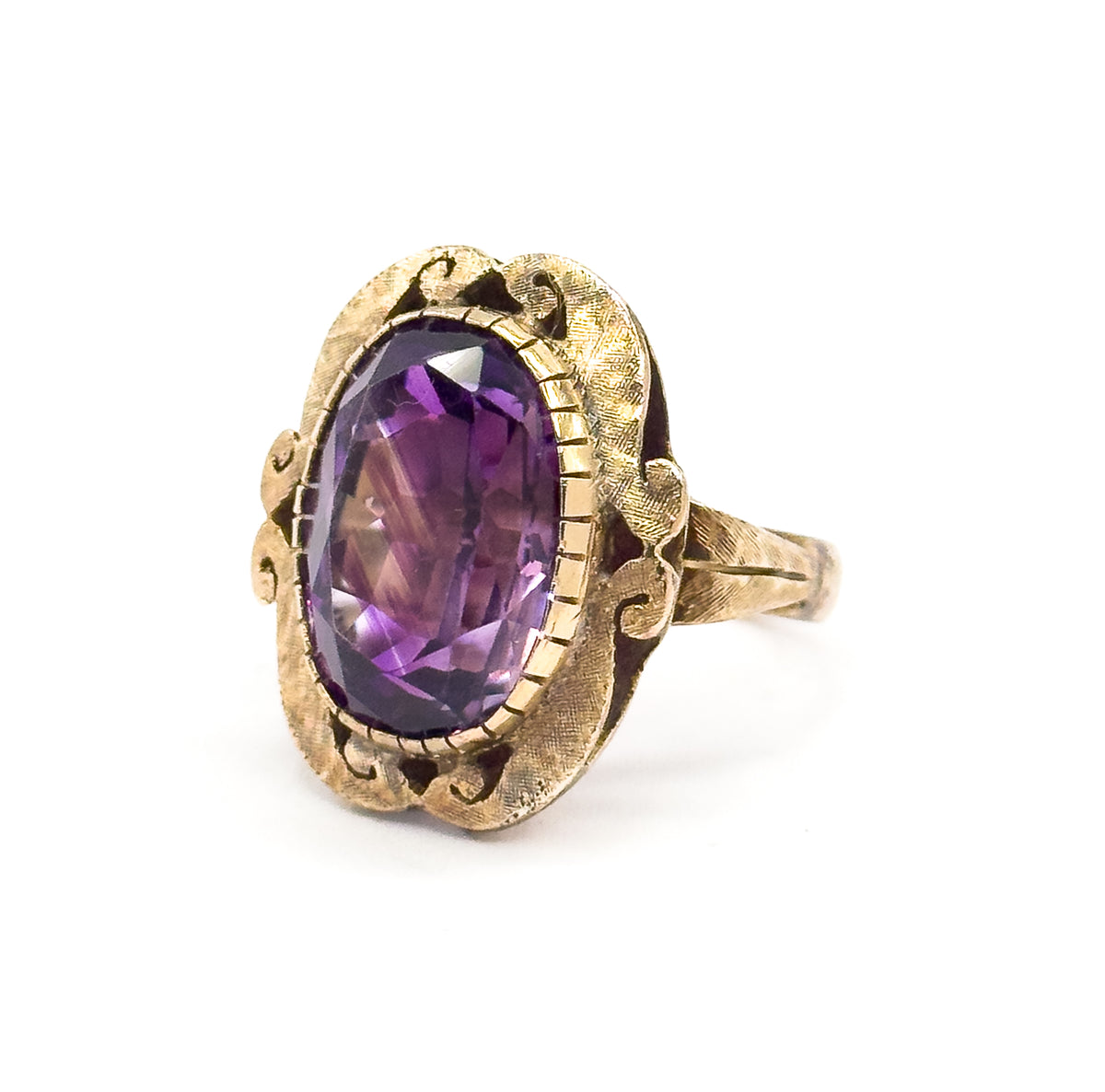Angled from the front left, the oval amethyst fills the ridged bezel with deep purple and paler flashes through its facets, framed by the matte textured face and scrolled shoulders.