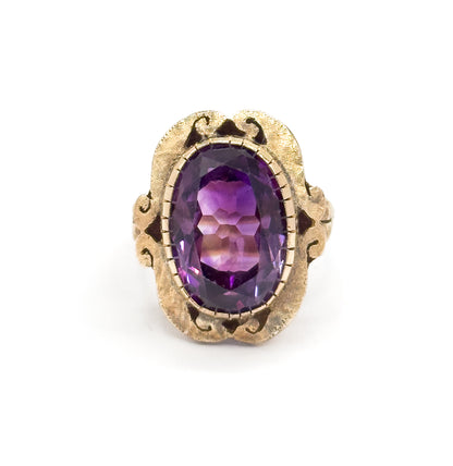 Straight-on from the front, the oval amethyst sits centred in its ridged bezel within a symmetrical textured face whose scrolled cutouts flare evenly to either side above the shank.