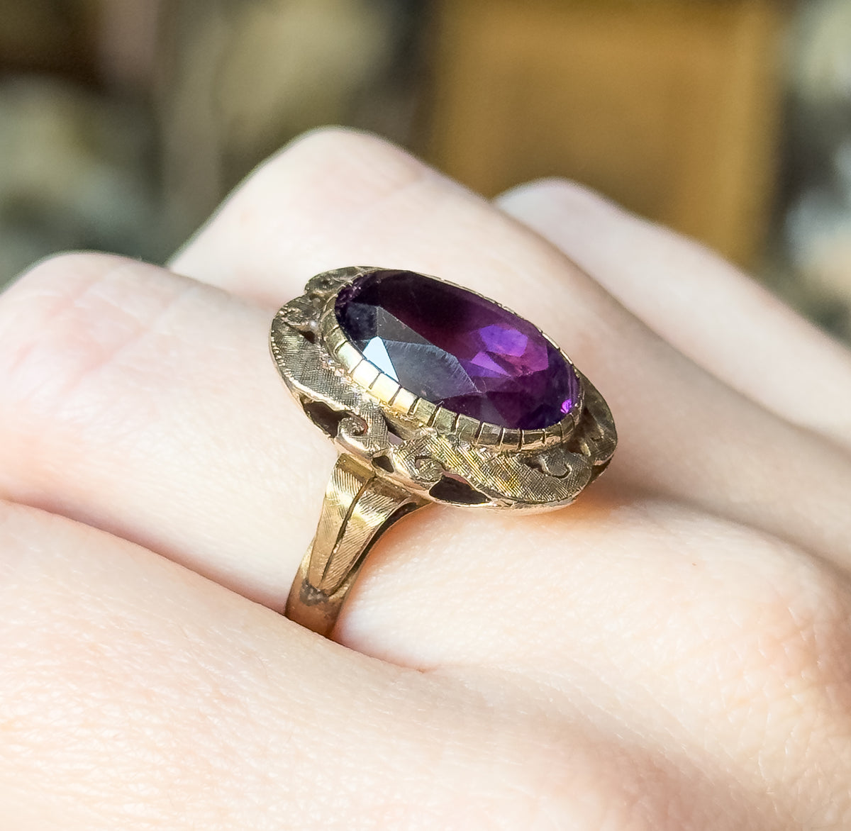 Mid-Century amethyst ring in 14K yellow gold, shown on a finger, its oval purple stone in a textured bezel with cutout detailing above a tapering shank.