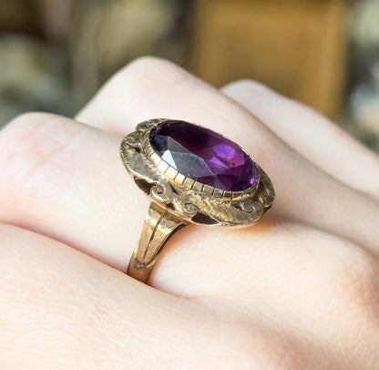 Mid-Century amethyst ring in 14K yellow gold, shown on a finger, its oval purple stone in a textured bezel with cutout detailing above a tapering shank.