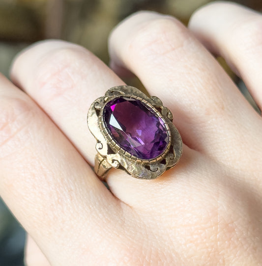 A closer angled view shown on the finger, the oval amethyst catching light across its facets within the scalloped, matte-textured bezel with scrolled cutouts at each side.