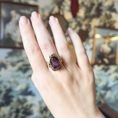 Shown at a distance on the ring finger of a whole hand, giving a sense of the face's scale against the knuckles, with framed pictures blurred behind.