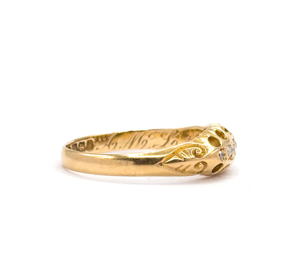 Low side angle on white showing the ring's shallow profile, the scrolled shoulder rising to the setting where two diamonds catch the light, and cursive interior engraving along the inner shank.