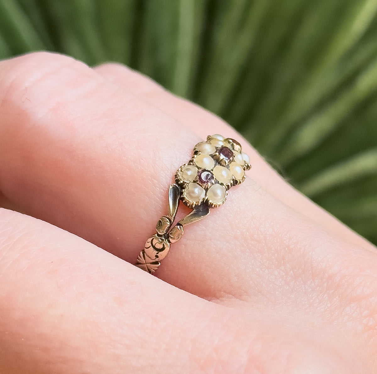Shown on a finger against blurred green foliage, the close view giving a sense of its small scale, with the leaf-carved shoulders and floral motifs on the shank clearly visible.