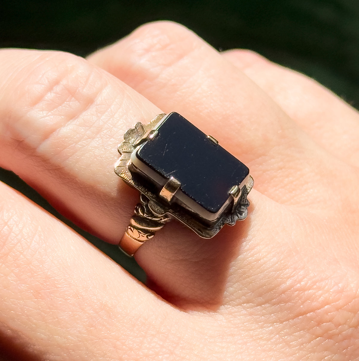 Shown on a finger in daylight, the rectangular sardonyx tablet and its prongs filling the finger's width, with the patterned shoulders and engraved shank visible alongside the skin for scale.