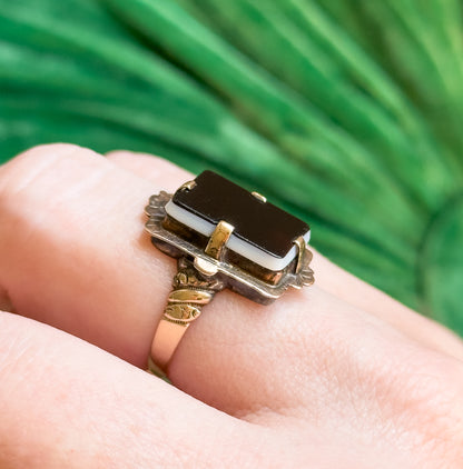 Close three-quarter view on a finger against blurred green foliage, the black-over-white layering of the sardonyx clear in sunlight above the scalloped mount and engraved shoulder.