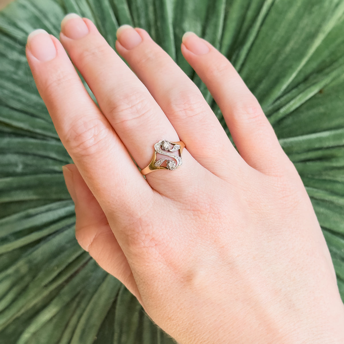 Shown on a finger against a green palm leaf, the ring's small size is clear, its white gold "S" face with two diamonds sitting low between the yellow gold shoulders.