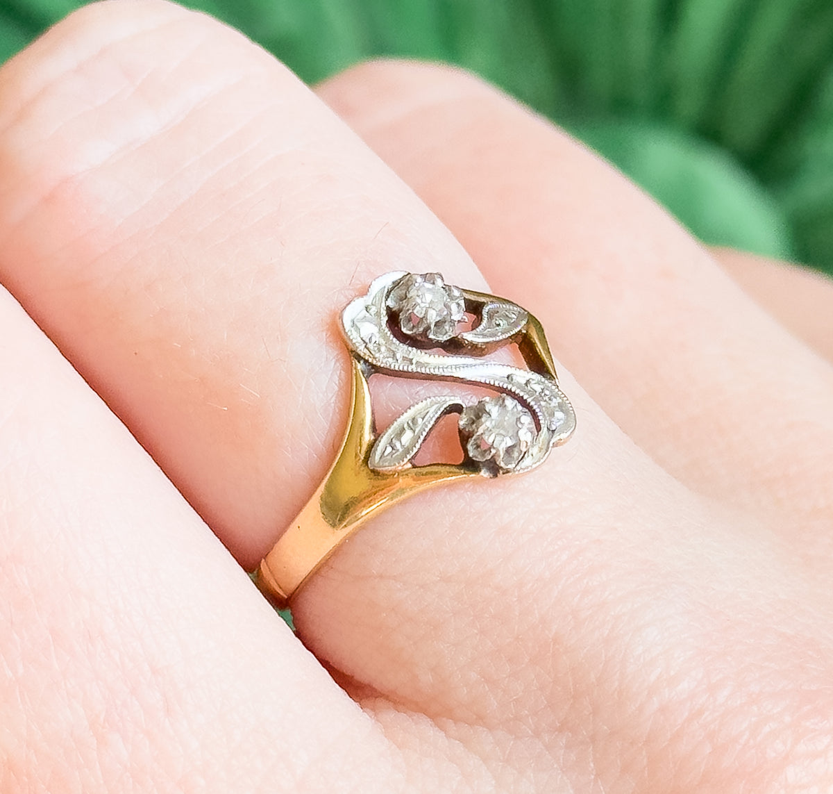 Close view on the finger from above, the two prong-set diamonds and milgrain-edged openwork of the white gold "S" face filling the frame against the yellow gold shank.