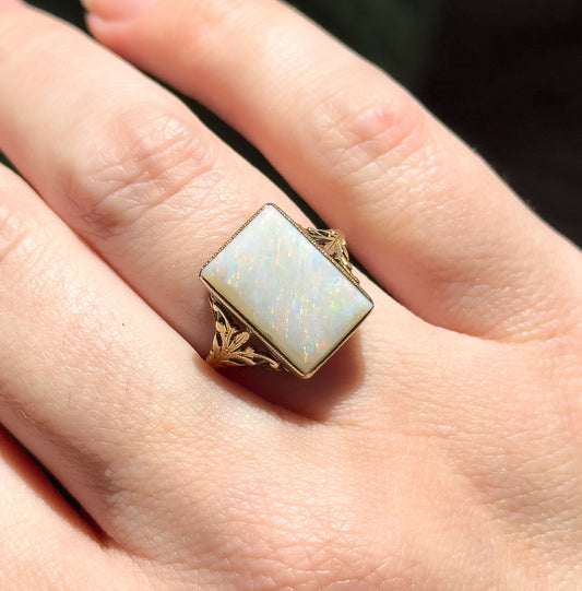 Shown on a finger in daylight, the rectangular opal fills the finger's width with red flashes across its white body, between leafy openwork gold shoulders.