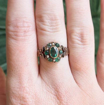 Shown on a finger against a green background, the cluster face sitting low and spanning about half the finger's width, with scrolled shoulders visible either side.