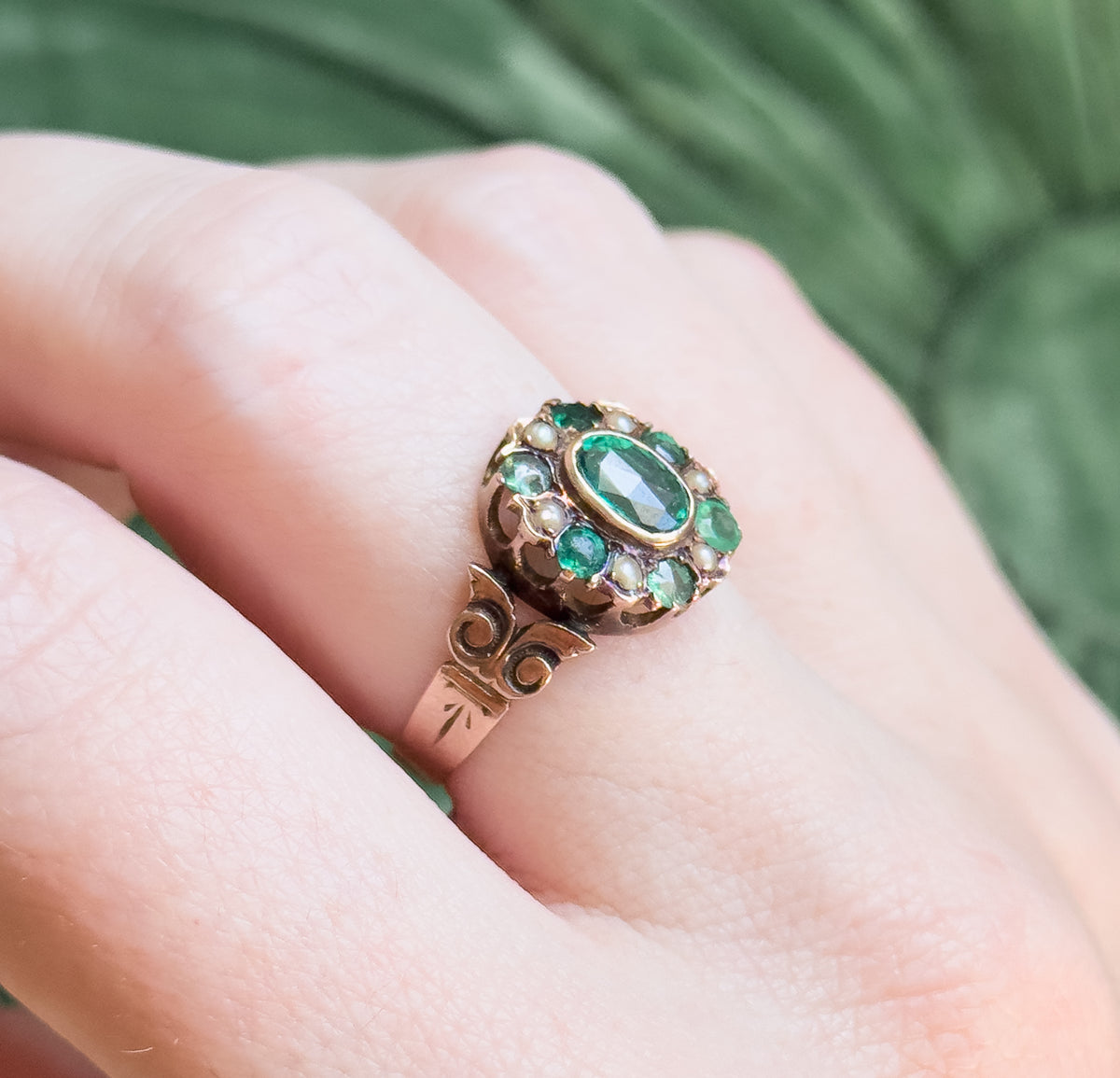 Close view on the hand from the side, the green oval and its halo of small green stones and seed pearls catching the light above the scrolled shoulder and engraved shank.
