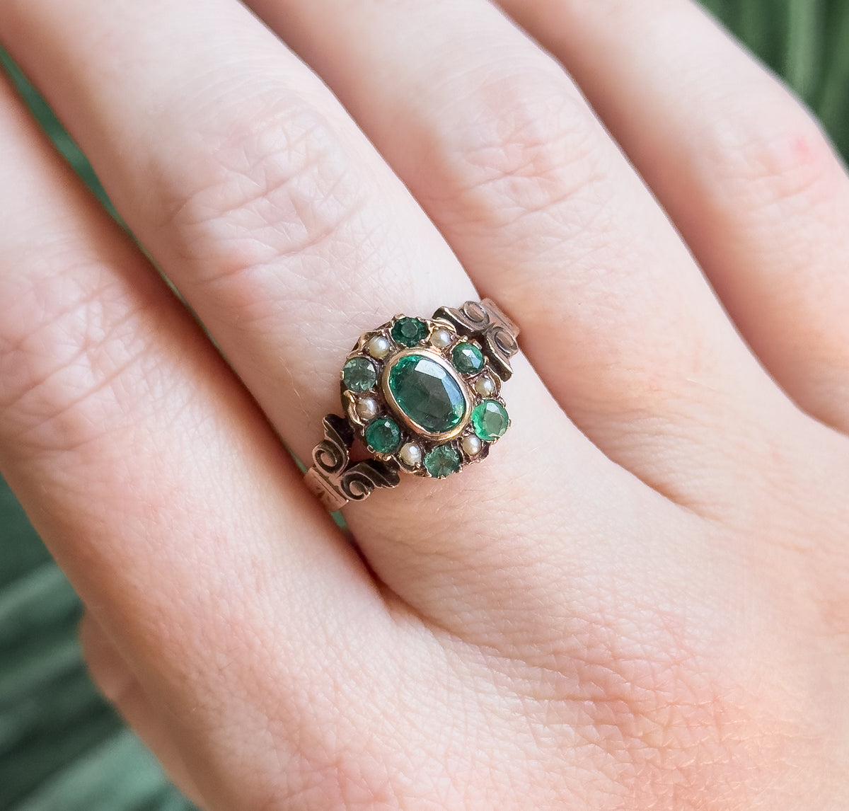 Looking down onto the hand from above, the bezel rim around the green oval is clear, with small green stones and seed pearls alternating around it and the shoulders curling away below.