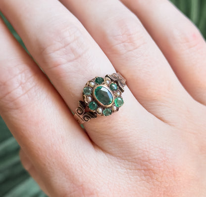 Looking down onto the hand from above, the bezel rim around the green oval is clear, with small green stones and seed pearls alternating around it and the shoulders curling away below.