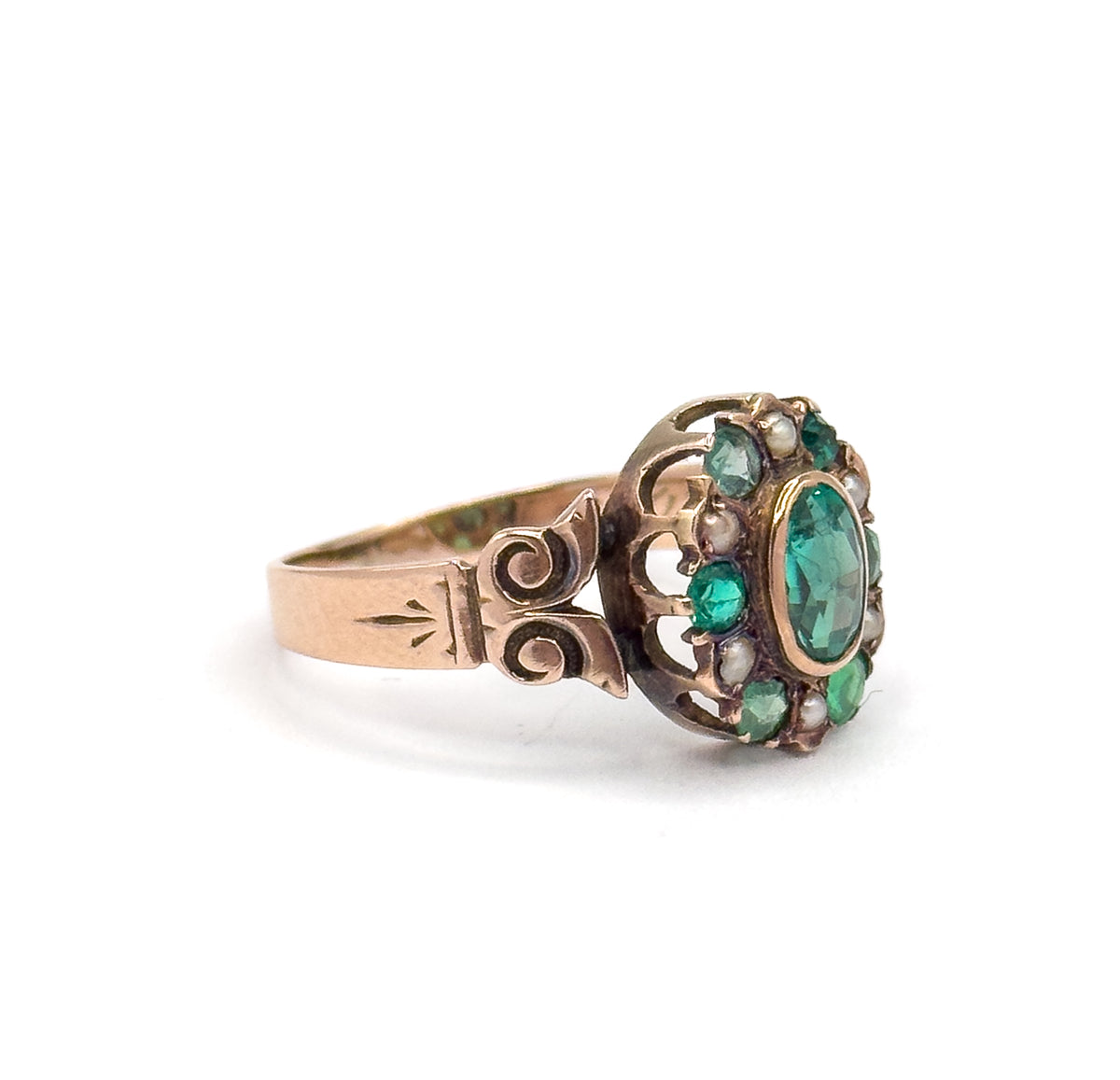 Side profile against white showing how low the bezel sits above the finger line, with the scrolled shoulder and arrow-like engraving leading up to the green stones and seed pearls.