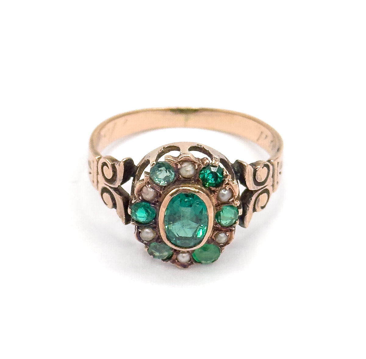 Head-on view of the face against white, the bezel-set green oval ringed by alternating smaller green stones and seed pearls, with open scrollwork shoulders flanking it and the plain band rising behind.