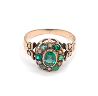 Head-on view of the face against white, the bezel-set green oval ringed by alternating smaller green stones and seed pearls, with open scrollwork shoulders flanking it and the plain band rising behind.