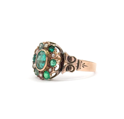 Victorian green stone and seed pearl gold ring shown in profile against white, the bezel-set oval centre ringed by smaller green stones and pearls above scrolled shoulders and a tapering shank.