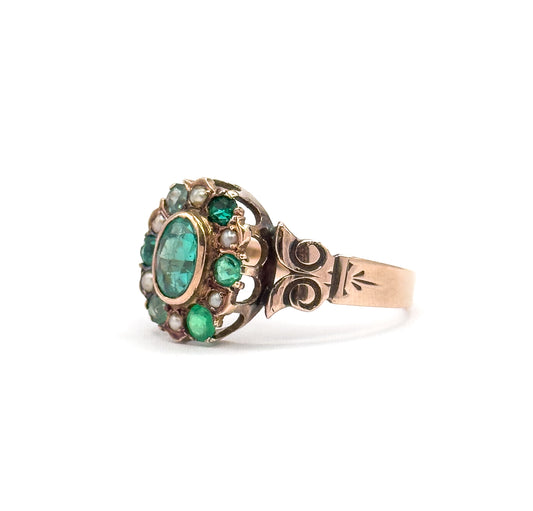 Victorian green stone and seed pearl gold ring shown in profile against white, the bezel-set oval centre ringed by smaller green stones and pearls above scrolled shoulders and a tapering shank.