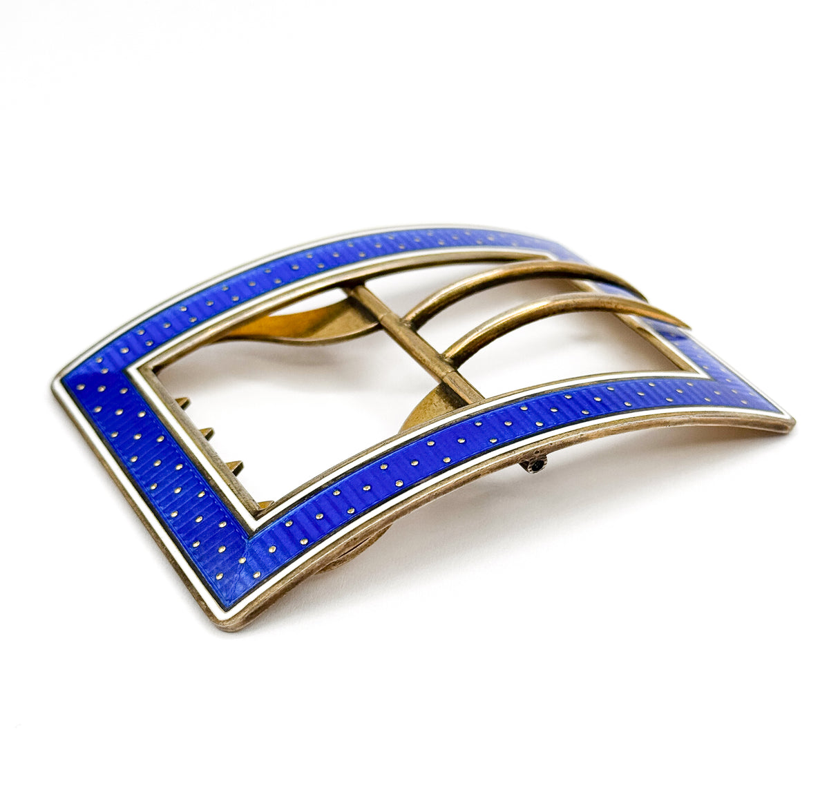 Low three-quarter view that reveals the buckle's gentle lengthwise curve, the blue and white enamel bands rising over the frame edge while the twin tongues and notched bar sit in shadow beneath.