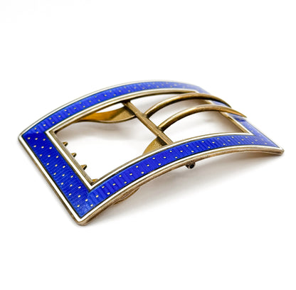 Low three-quarter view that reveals the buckle's gentle lengthwise curve, the blue and white enamel bands rising over the frame edge while the twin tongues and notched bar sit in shadow beneath.