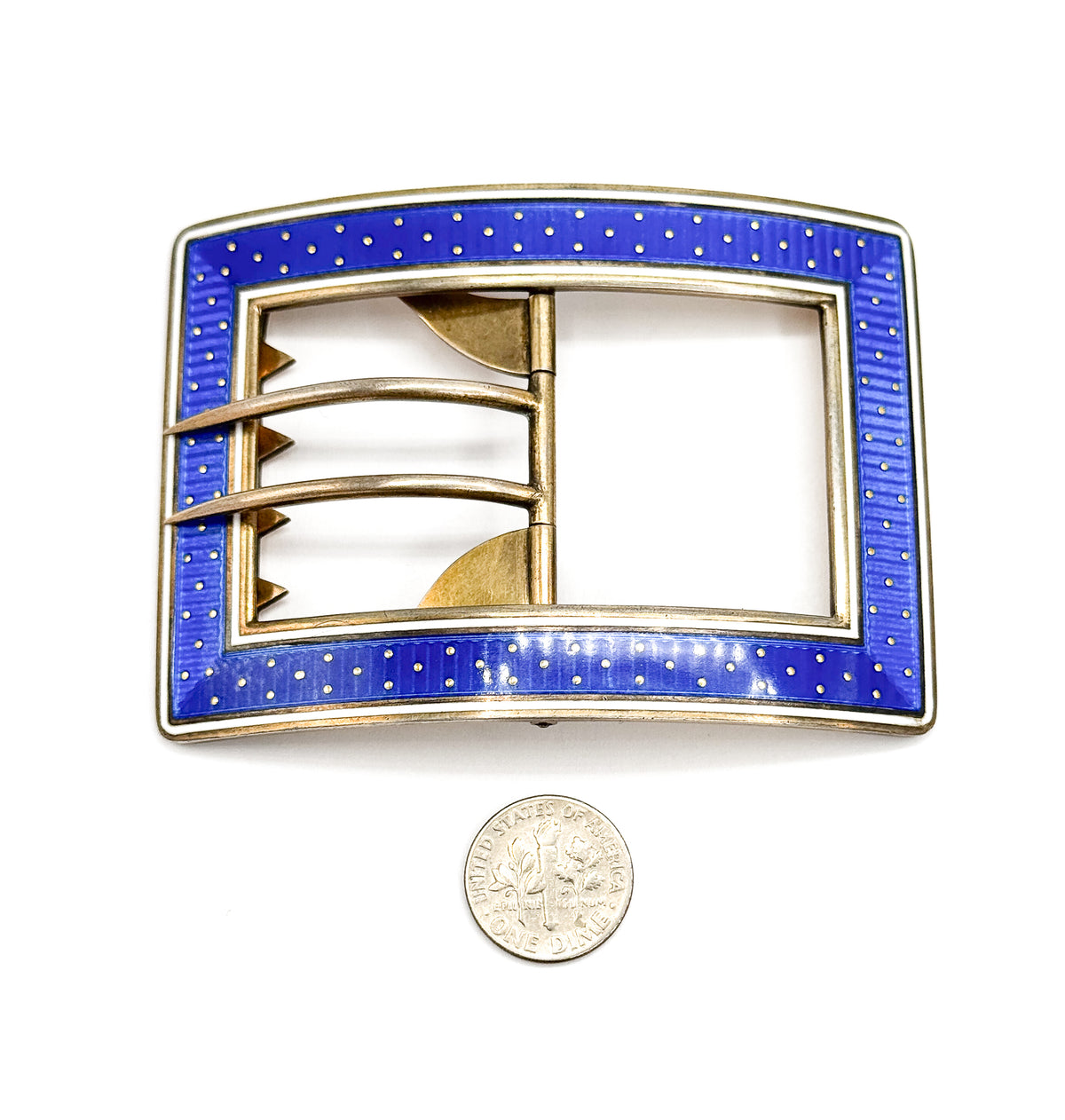 The buckle flat and face-up beside a dime, which shows its size, with the blue enamel band of gold dots framing the open centre, hinged tongue and notched bars.