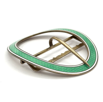 Angled three-quarter view from the back, the single prong lying diagonally across the open oval and the scalloped crossbar and hinge visible against the green and white enamel rim.
