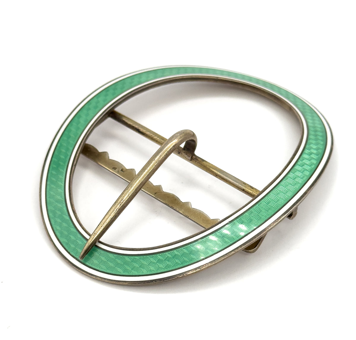 Tilted rear view with the buckle lying almost flat, the prong crossing the open centre and small fittings standing proud at the right edge, the green and white enamel rim encircling it all.