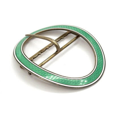 Angled three-quarter view of the whole oval from above, the mint green enamel band with its white border widening toward the front and the prong hooked over the straight bar across the open centre.