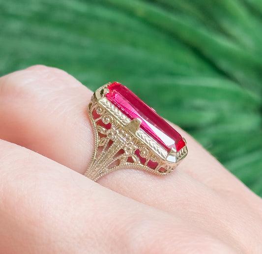 Shown on a finger at a low angle against blurred green foliage, the elongated pinkish-red stone and its scrolled filigree gallery rising above the narrow shank.