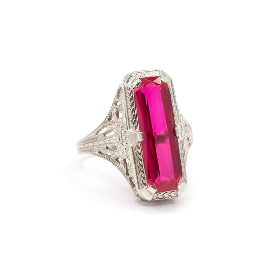 An Art Deco synthetic ruby filigree ring in 14K white gold, shown upright against white, the long pinkish-red stone prong-set in lacy openwork above a slender shank.