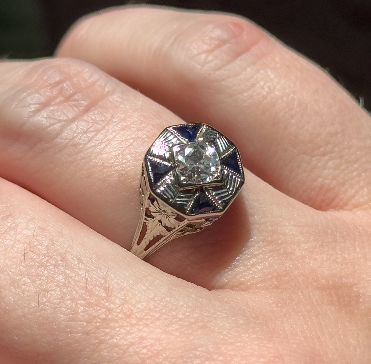 Shown on a finger in daylight, the octagonal face raised above the knuckle so the etched shoulders and pierced filigree gallery along the tapering shank are visible.