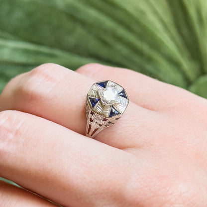 Shown on a finger against blurred green foliage, the octagonal face angled towards the camera so the four triangular blue stones and etched rays around the central diamond read clearly.